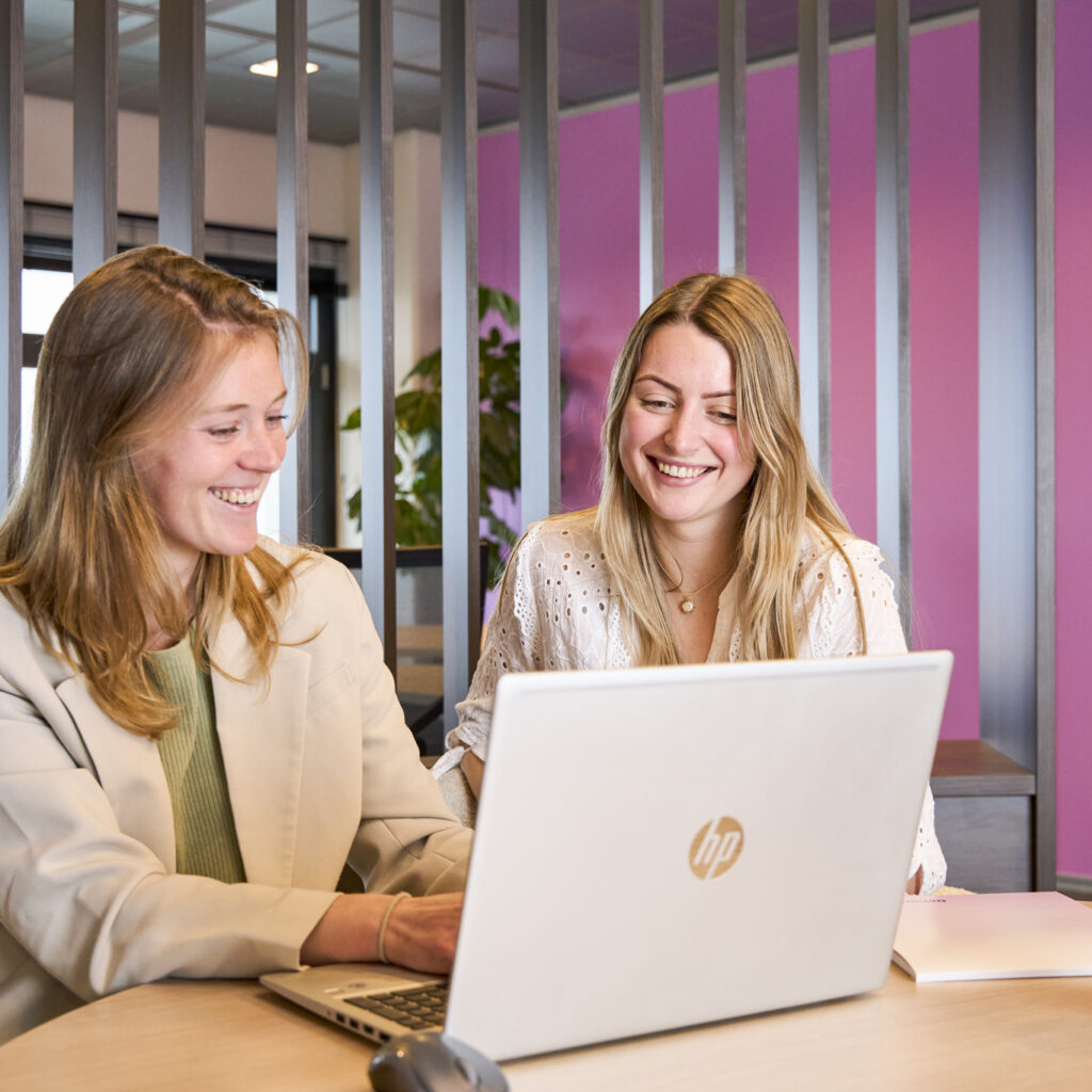 Twee vrouwelijke collega's bij Déhora die samenwerken en lachen achter een laptop in een modern kantoor.